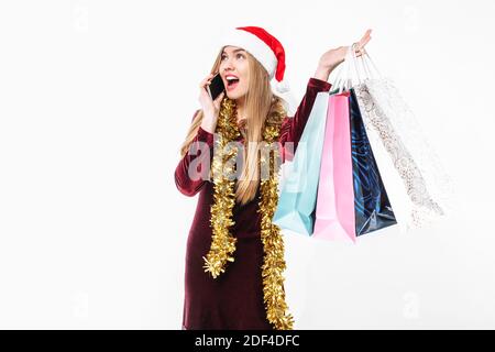 Belle femme en chapeau de père noël, avec des sacs de noël en main, parlant sur un smartphone. Sur fond blanc. Banque D'Images