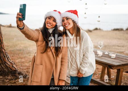 Jeunes filles souriantes de course mixte faisant un autoportrait À l'extérieur de votre téléphone, port du chapeau du Père Noël en mer jardin du complexe chaleureux Banque D'Images