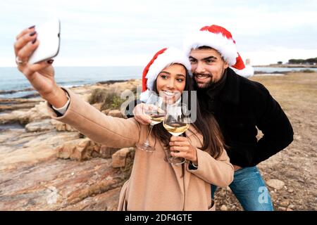 Jeune beau couple multiracial faisant un autoportrait à l'extérieur station balnéaire portant un chapeau de Père Noël tenant des verres à vin blancs Célébration de Noël Banque D'Images