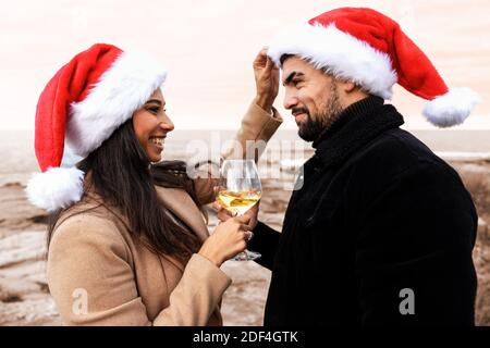 Jeune beau couple multiracial qui boit du vin à l'extérieur du Père Noël chapeau célébrant les vacances dans la station balnéaire en hiver - Beau heureux Banque D'Images