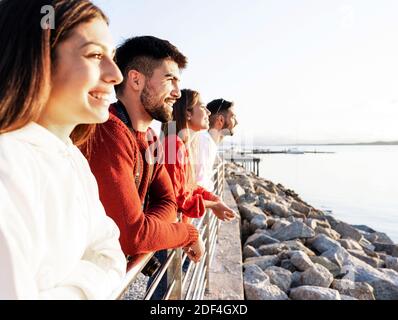 Jeunes gens heureux et beaux penchée sur une rambarde en métal à le coucher de soleil sur l'eau en mer hiver station de vacances - Groupe d'amis confiants à regarder Banque D'Images