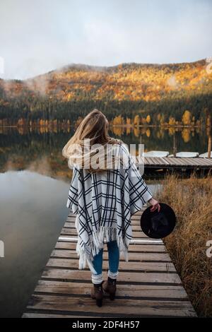 Jeune femme blonde se tenant sur la jetée en bois près de la forêt d'automne. Voyage, concept de style de vie. Banque D'Images