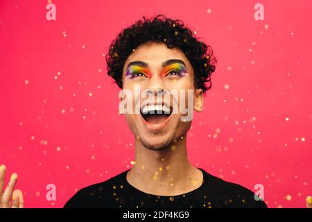 Homme androgyne avec maquillage pour les yeux arc-en-ciel, qui jette les paillettes et rit. gays excités avec des paillettes volant sur fond rouge. Banque D'Images