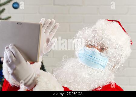 Homme en costume du Père noël et masque de grippe avec un ordinateur tablette. Félicitations du père noël via tablette. Banque D'Images