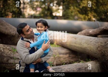 Un père de brunette indien avec son bébé garçon sur ses genoux en hiver après-midi assis sur des rondins en bois en arrière-plan de la forêt. Banque D'Images