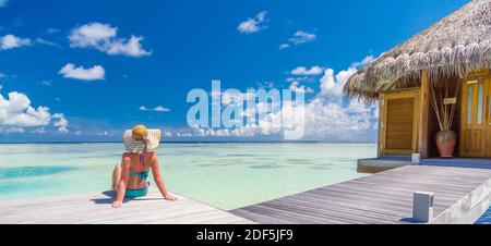 Bonne jeune femme assise sur une villa aquatique dans un complexe de luxe. Profitez de la vie, détendez-vous dans une ambiance d'été. Femme en chapeau regardant le style de vie de loisir de mer Banque D'Images