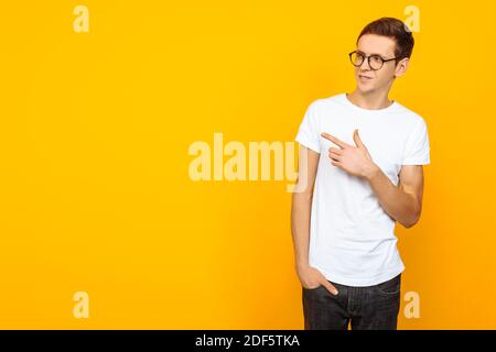 Homme attrayant, portant des lunettes, en t-shirt blanc vierge, pointant avec deux doigts sur le côté, pointant vers l'espace vide, sur fond jaune Banque D'Images