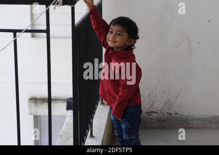 Portrait d'Indien mignon petit gai brune petit garçon tamoul porter une chemise rouge vif tout en étant debout sur un balcon à l'intérieur un fond urbain blanc Banque D'Images