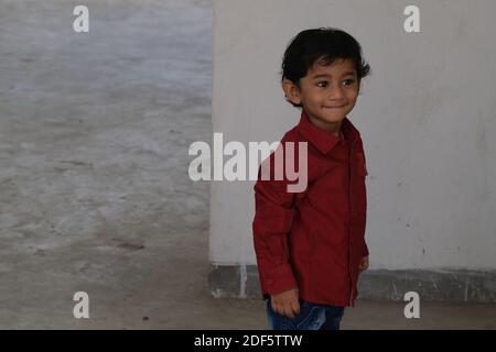 Portrait d'Indien mignon petit gai brune petit garçon tamoul porter une chemise rouge vif tout en étant debout sur un balcon à l'intérieur un fond urbain blanc Banque D'Images