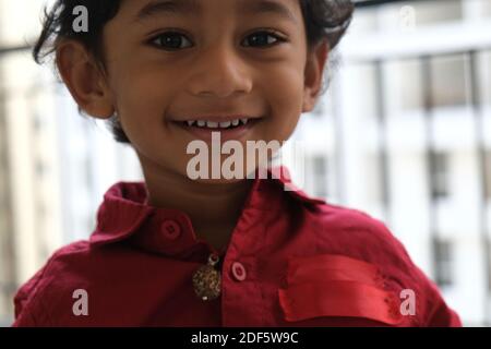 Portrait d'Indien mignon petit gai brune petit garçon tamoul porter une chemise rouge vif tout en étant debout sur un balcon à l'intérieur un fond urbain blanc Banque D'Images