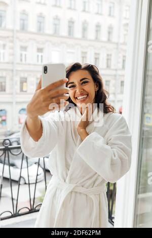 Bonne femme prenant le selfie sur un balcon. Jeune femme en peignoir tenant un smartphone et faisant des photos. Banque D'Images