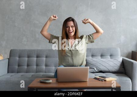 Une jeune femme surjoyée s'assoit sur un canapé à la réception du triomphe à la maison e-mail agréable ou lire les bonnes nouvelles sur ordinateur portable en ligne Banque D'Images