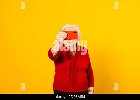 Portrait studio d'une femme âgée portant une chemise rouge sur fond jaune et passer un appel vidéo avec un téléphone mobile rouge Banque D'Images