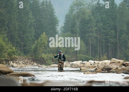 Pêcheur dans des vêtements spéciaux et des bottes utilisant la tige professionnelle pour attraper le poisson à la rivière. Homme debout dans l'eau parmi les montagnes vertes. Con. Relaxation Banque D'Images