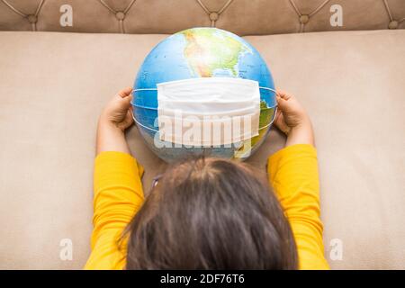 La petite fille tient le globe terrestre entre ses mains. Masque chirurgical blanc sur le globe terrestre. Concept de pandémie. Concept de séjour à la maison. Banque D'Images