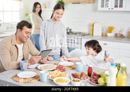 Mari latin d'âge moyen utilisant un Tablet pc, regardant les informations, attendant le dîner tandis que ses enfants et sa femme servent une table dans la cuisine à la maison Banque D'Images