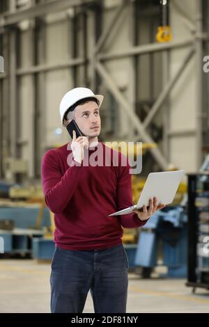 Portrait d'un directeur d'usine masculin dans un casque blanc et un chandail rouge tenant un ordinateur portable et un téléphone portable. Contrôler le processus de travail au fabricant de l'hélicoptère. . Photo de haute qualité Banque D'Images