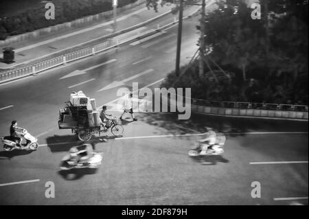 Man riding tricycle transportant une montagne de vieux meubles et du personnel. Au milieu d'un Carrefour en Chine. Photo en noir et blanc. Banque D'Images