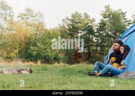 Beau couple assis à l'entrée de la tente avec tasses dans leurs mains et regardant sur le feu de camp Banque D'Images