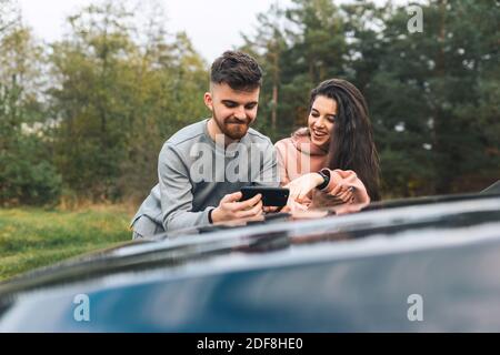 Le jeune couple regarde quelque chose de gai dans le smartphone, debout sur le capot de la voiture Banque D'Images