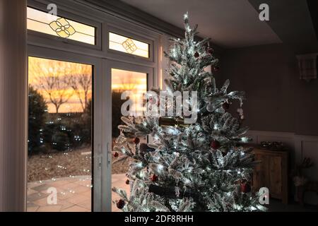 Peu profonde attention sur les décorations sur un joli arbre de Noël givré atifical avec des lumières de fée devant les portes de patio pendant un coucher de soleil doré. Banque D'Images