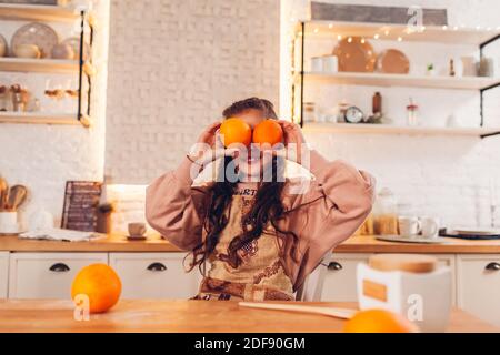 Petite fille jouant avec deux fruits oranges au lieu des yeux sur la cuisine. Enfant s'amusant, cuisant en portant un tablier à la maison Banque D'Images