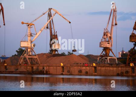 des grues de manutention en vrac à lubrification de niveau chargent du sable sur une barge de cargaison sèche dans un port de rivière à la lumière du soir Banque D'Images