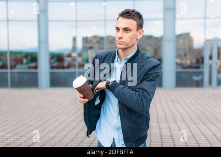 Un homme avec une tasse de papier de café, passe à travers la ville, un beau gars en vêtements élégants, sort son téléphone de sa poche, le temps est bon Banque D'Images