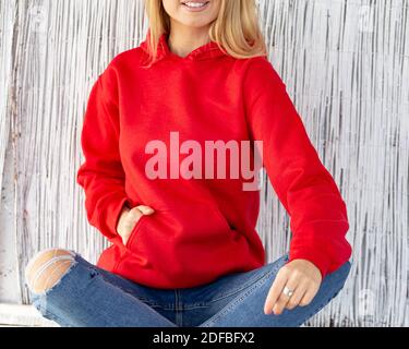 Une femme blonde non reconnaissable est assise devant l'appareil photo. Elle porte un pull à capuche rouge de Noël avec un endroit pour l'incriion. Tenue de fête pour mockup avec espace pour les copies. Banque D'Images