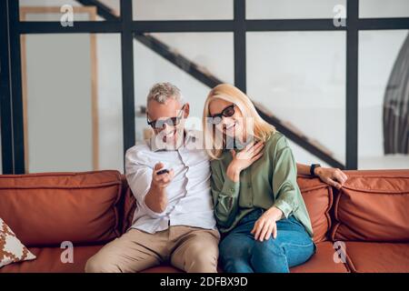 Couple d'âge mûr en lunettes de soleil assis sur le canapé et sensation joyeux Banque D'Images