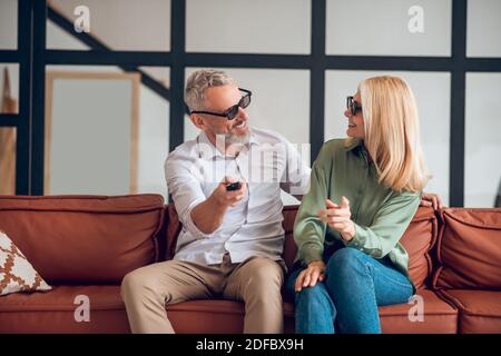 Couple d'âge mûr en lunettes de soleil assis sur le canapé et commutation sur le téléviseur Banque D'Images