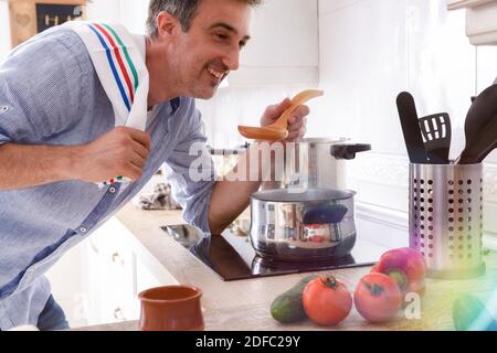 Homme souriant testant la soupe de légumes avec une cuillère en bois dans la cuisine Banque D'Images