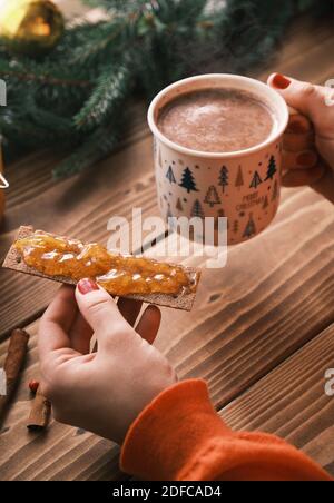 Les mains des femmes tenant la confiture sur un pain croustillants de près avec une tasse de chocolat chaud. Petit déjeuner de Noël Banque D'Images