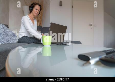 Femme, mi-50, travaille depuis la maison, avec un ordinateur portable et communique par micro-casque avec ses collègues, son bureau à domicile, sur le canapé, Banque D'Images