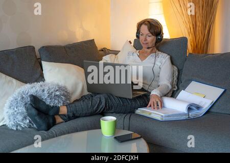 Femme, mi-50, travaille depuis la maison, avec un ordinateur portable et communique par micro-casque avec ses collègues, son bureau à domicile, sur le canapé, Banque D'Images