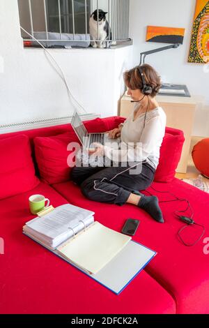 Femme, mi-50, travaille depuis la maison, avec un ordinateur portable et communique par micro-casque avec ses collègues, son bureau à domicile, sur le canapé, Banque D'Images