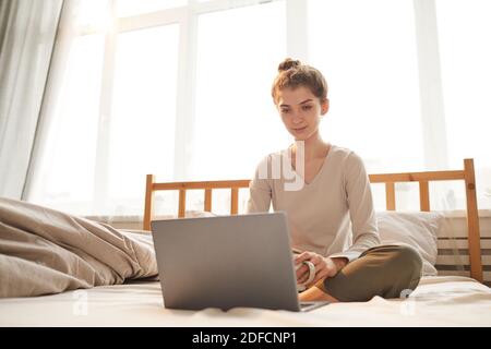 Portrait d'une jeune femme regardant l'écran d'un ordinateur portable pendant assis sur le lit le matin dans la chambre Banque D'Images