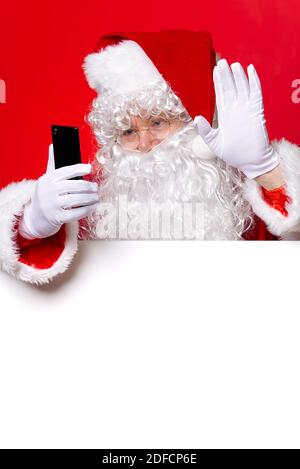 Le Père Noël tient le téléphone portable en main tout en se tenant derrière le tableau blanc vierge. Espace de copie. Envoi de SMS au Père Noël. Banque D'Images