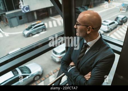 Vue de dessus. Un homme d'affaires chauve et contemplatif en costume et en lunettes qui donne loin dans la fenêtre ou l'ascenseur en verre avec une vue animée sur la rue. Pensif Banque D'Images