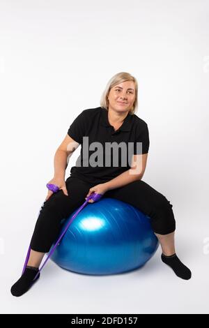 Femme âgée faisant des exercices sur une balle de fitness et avec une bande élastique. Photo sur fond blanc. Banque D'Images