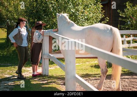 Bonne vie de ferme. Mère et fille dans un stand de vêtements vintage près du paddock avec un beau cheval blanc. La fille est un cheval de course. Famille à Banque D'Images