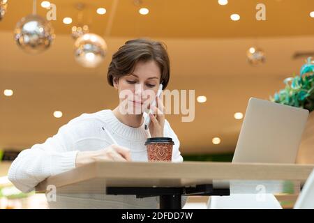 le programmeur est assis dans un café à un ordinateur portable, écrit dans un bloc-notes, une photo de dessous. Brunette en chandail blanc, concept indépendant Banque D'Images