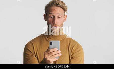 Surpris homme barbu porter turtleneck regarder stupéfait bavarder avec des amis sur smartphone isolé sur fond blanc. Wow visage Banque D'Images