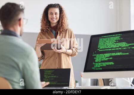 Une jeune femme heureuse avec une tablette numérique en consultation avec son collègue pendant qu'il travaille sur un ordinateur avec un logiciel Banque D'Images