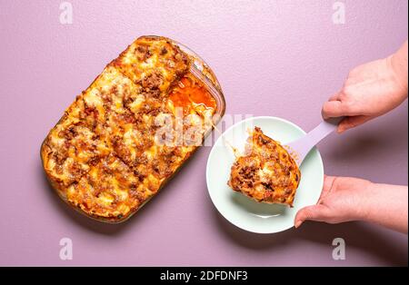 Vue ci-dessus avec les mains de femme servant sur une assiette une portion de lasagnes fraîchement cuites. Délicieux lasagne maison vue sur un fond violet. Banque D'Images