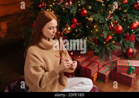 Photo en grand angle d'une jeune femme rêveuse qui écrit la liste de souhaits pour Nouvel an sur fond d'arbre de Noël Banque D'Images