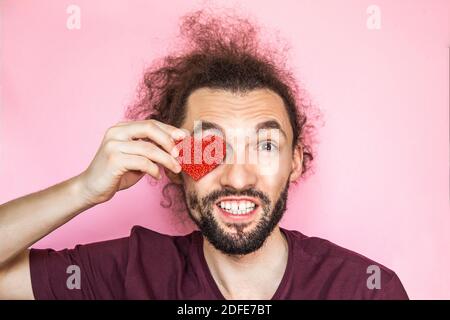 Portrait en gros plan de l'homme beau et émotionnel barbu oeil fermé par coeur rouge, isolé sur rose Banque D'Images