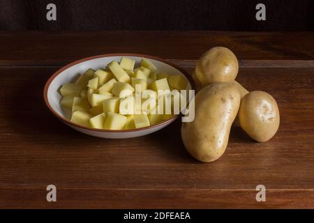 Un bol est rempli de pommes de terre coupées en dés à côté de pommes de terre entières dans leur peau sur une table en bois de châtaignier dans un intérieur de maison. Ingrédients naturels et biologiques. Banque D'Images