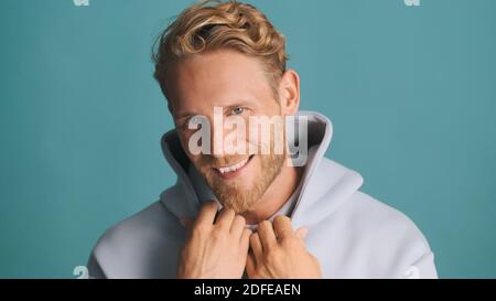 Beau sourire barbu homme en sweat à capuche regardant avec joie dans l'appareil photo et souriant isolé sur fond bleu. Modèle mâle Banque D'Images