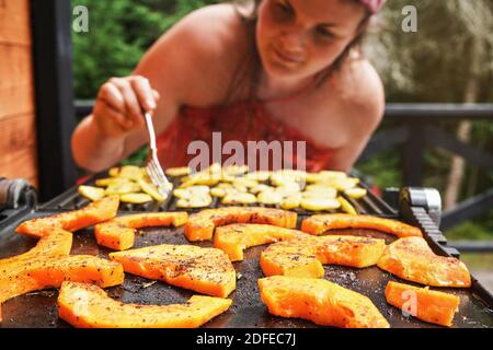 Morceaux de courge de noyer cendré grillés sur le gril électrique, se concentrer sur les légumes orange vif assaisonnés d'épices, flou jeune femme en arrière-plan Banque D'Images